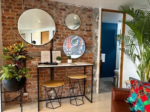 Une pièce avec un mur en briques, un miroir rond, une table avec deux tabourets, une plante en pot et un coussin coloré.