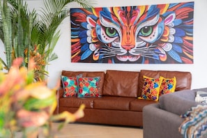 Een bruine leren bank met kleurrijke kussens met bloemenprint, een levendig abstract schilderij van een kattengezicht aan de muur, en een potplant aan de linkerkant.