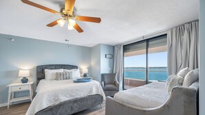 Une chambre à coucher comprenant un lit, une chaise, une table de chevet, une lampe, un ventilateur de plafond et une fenêtre donnant sur la mer.