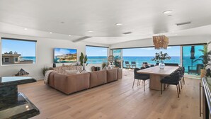 A modern living room with a large sofa, dining table, and chairs, large windows with ocean views, and a hanging light fixture. - Beachfront Boho Dream Penthouse. Walk 2 Everything!   Newly designed  SLEEPS 10! (Oceanside)