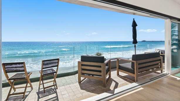 A balcony with wooden chairs and a table, overlooking a beach with waves and a clear sky. - Beachfront Boho Dream Penthouse. Walk 2 Everything!   Newly designed  SLEEPS 10! (Oceanside)