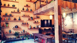 Een gezellig interieur met houten planken waarop diverse keramiek en planten staan, een houten bank en een eethoek met tafels en stoelen.