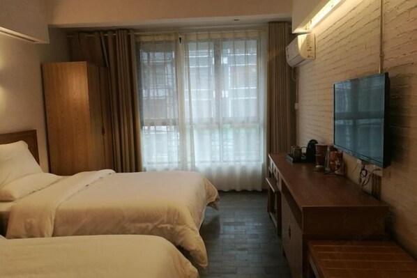 Quarto de hotel com duas camas, cômoda de madeira, televisão de tela plana e janela com cortinas translúcidas.