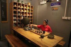 Une personne est assise à une table en bois, devant une machine à café, des tasses et un bol. Derrière elle, on aperçoit une étagère remplie de divers objets.