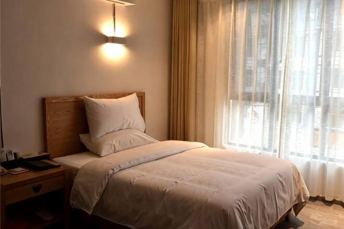 Ein ordentlich bezogenes Bett mit Holz-Kopfteil, ein Nachttisch mit Lampe und ein Fenster mit leichtem Vorhang.