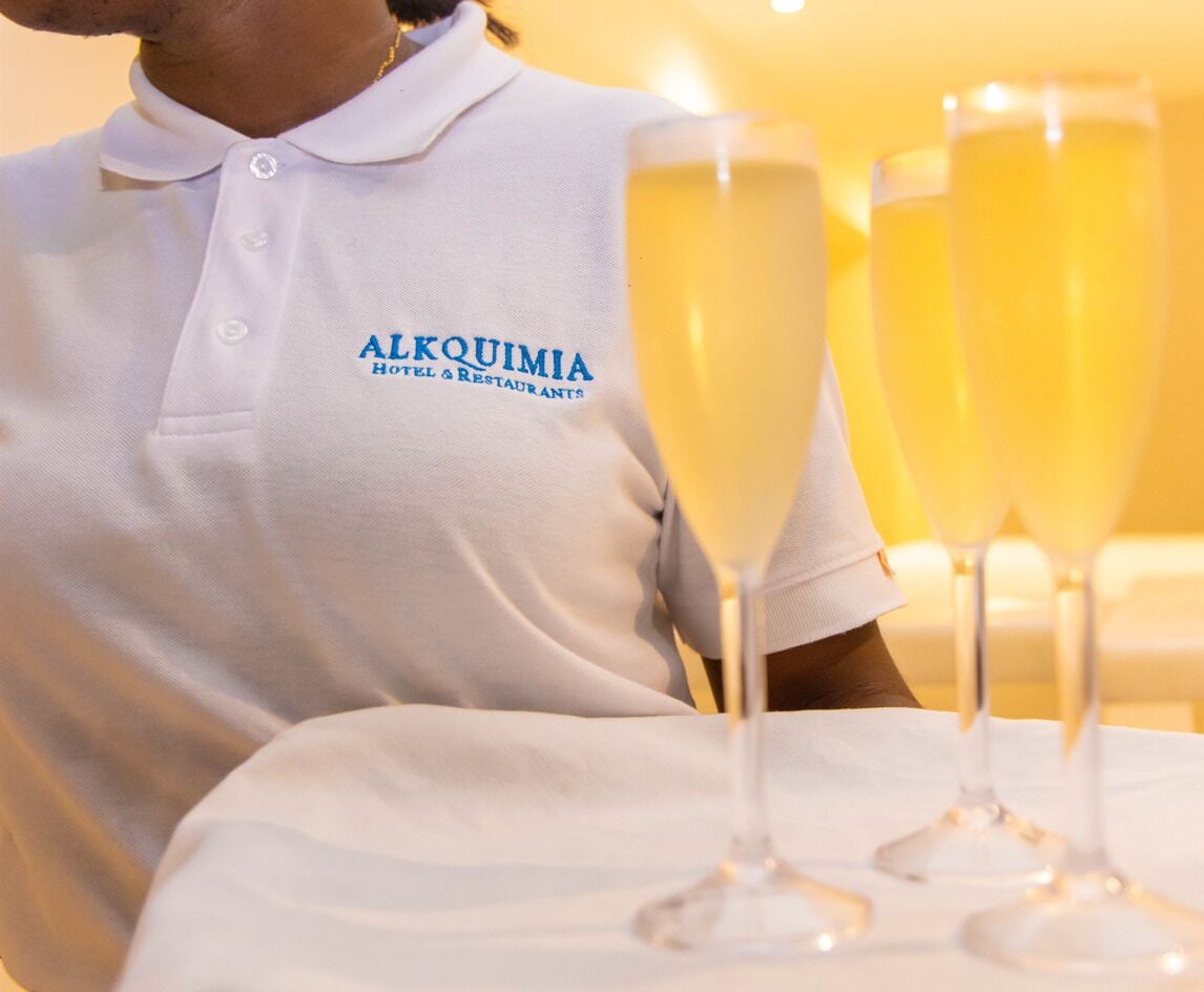 A person wearing a white polo shirt with "ALQUIMIA Hotel & Restaurants" printed on it, seated at a table with three champagne glasses.