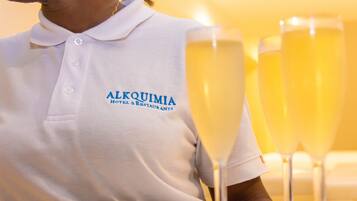 Uma pessoa vestindo camisa polo branca com a inscrição "ALQUIMIA Hotel & Restaurants", sentada à mesa com três taças de champanhe.