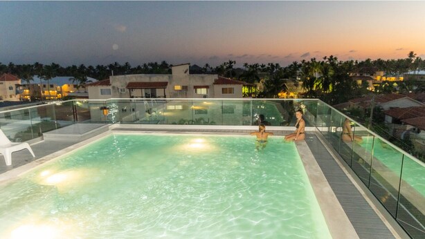 A rooftop infinity pool with clear water, surrounded by glass railings, and overlooking a residential area with palm trees and buildings. - Hotel Alkquimia (Bayahibe)