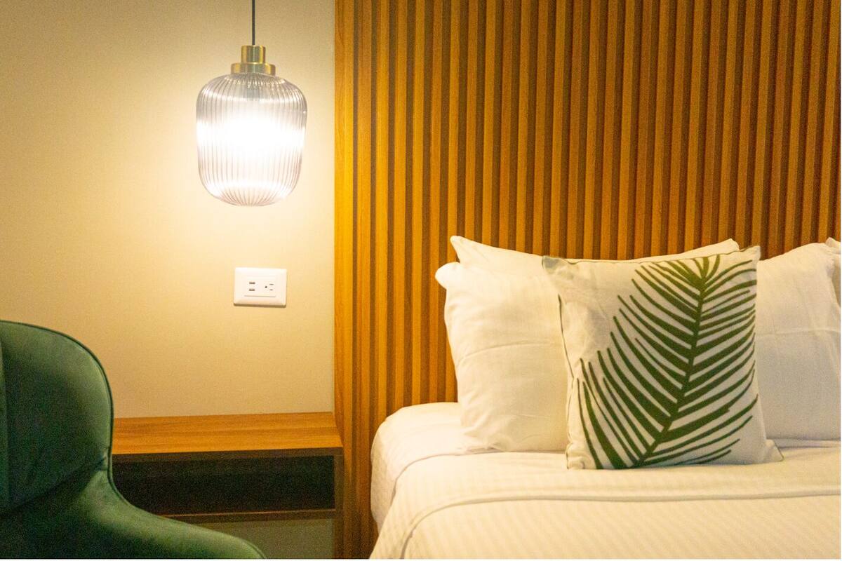 Une chambre d’hôtel avec un fauteuil recouvert d’un tissu vert, une table de chevet, un lit avec une literie blanche et un oreiller orné de motifs de feuilles de palmier, et une suspension lumineuse.