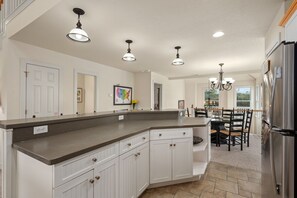 A kitchen with white cabinets, a granite countertop, and a stainless steel refrigerator. There are pendant lights and a chandelier. - Peace of Paradise: Private Pool, Hot Tub and Great Currituck Club amenities! (Corolla)