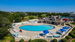 A large swimming pool with lounge chairs and umbrellas, surrounded by a wooden fence and greenery. - Peace of Paradise: Private Pool, Hot Tub and Great Currituck Club amenities! (Corolla)