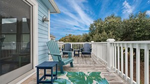 A wooden deck with a white railing, a blue chair, a small table, and a tropical leaf-patterned rug. - Peace of Paradise: Private Pool, Hot Tub and Great Currituck Club amenities! (Corolla)
