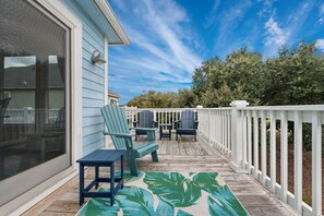 A wooden deck with a white railing, a blue chair, a small table, and a tropical leaf-patterned rug.