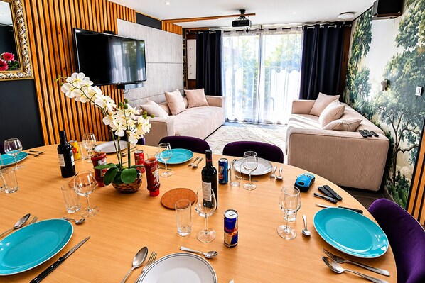 Un comedor con una mesa de madera dispuesta para una comida, un televisor montado en la pared y una sala de estar con sofás y vistas a zonas verdes.