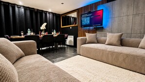 A modern living room with a sofa, a dining table with chairs, a TV mounted on the wall, and a decorative vase with flowers.