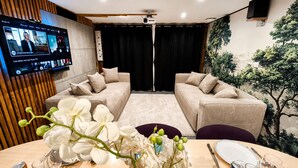 A modern living room with a flat-screen TV, two beige sofas, a wooden accent wall, and a dining area with a table set for two.