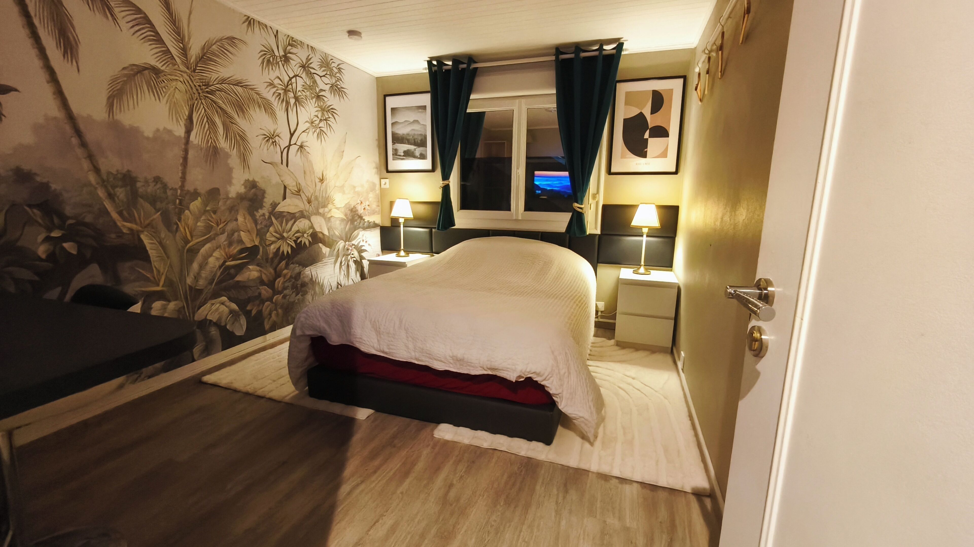 A hotel room with a bed, a desk, a nightstand, a lamp, a window with curtains, a wall mural, and a door.