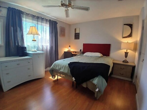 A bedroom with a bed, bedside tables, a desk, a chair, a ceiling fan, and a window with curtains.