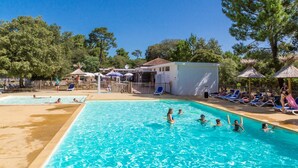 Una piscina con gente nadando y tomando el sol, rodeada de árboles y sombrillas.