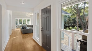 A spacious hallway with wooden flooring leading to a dark grey door, a living area with a grey sofa, and a view of a garden through large windows. - Great FAMILY home in Wild Dunes, Pond View, Modern, Minutes to Beach, Rent the Golf Cart! (Isle Of Palms)