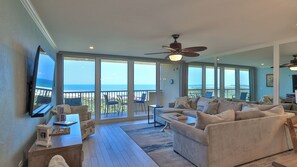 A spacious living room with a large window overlooking the ocean, a ceiling fan, and a cozy seating area.