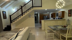 Un'ampia zona giorno con soffitto alto, una scala, un tavolo da pranzo con sedie e una cucina sullo sfondo.