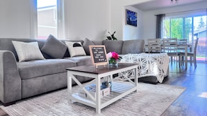 Un salon moderne avec un canapé gris, une table basse sur laquelle sont posés des jeux de société, un panneau d’accueil et un coin repas avec une table et des chaises en bois.