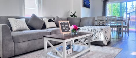 Una sala de estar moderna con un sofá gris, una mesa de centro con juegos de mesa, un letrero de bienvenida y una zona de comedor con una mesa y sillas de madera.