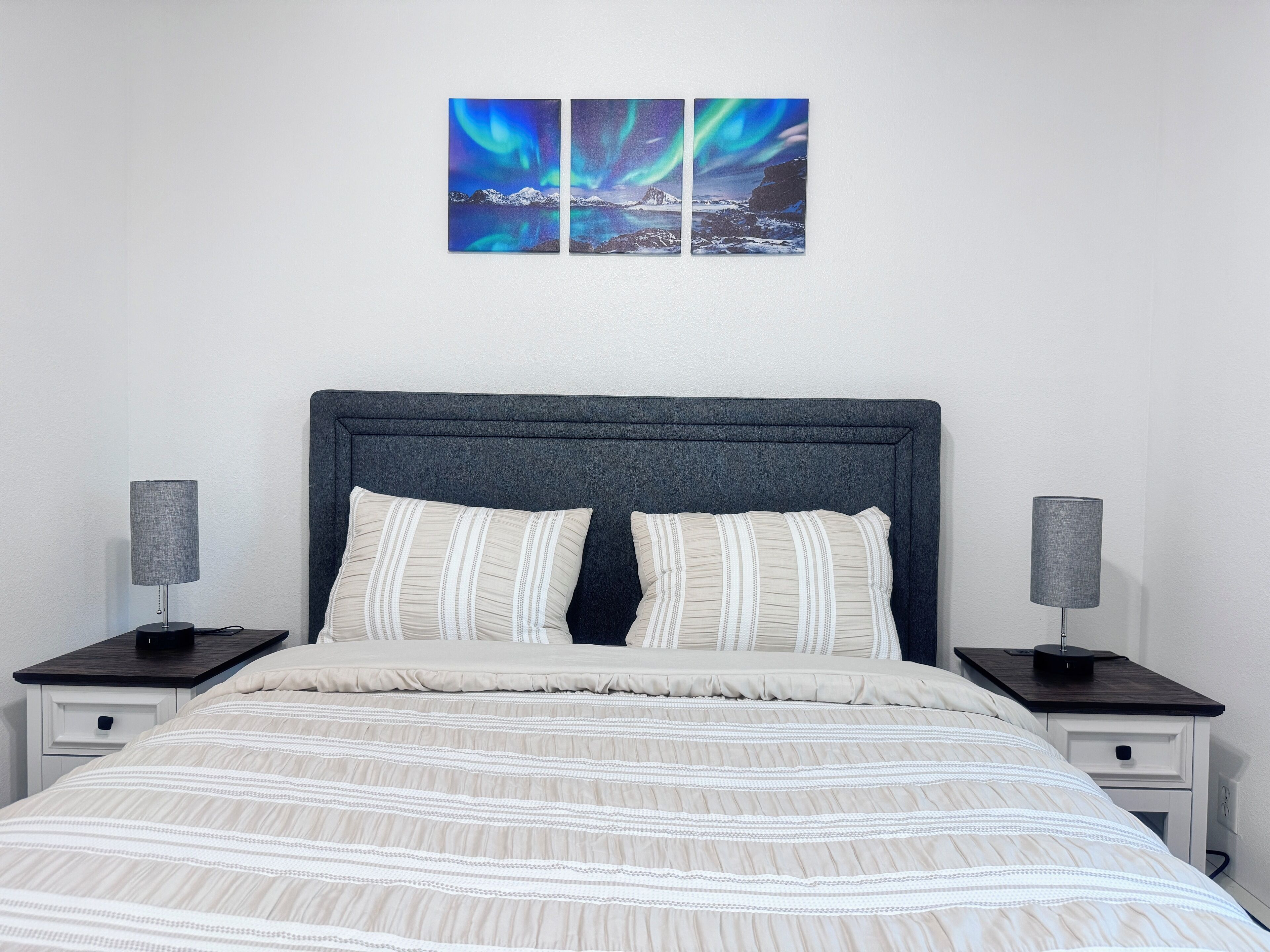A bedroom with a bed, two bedside tables, and a wall-mounted artwork of the Northern Lights.