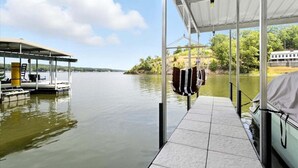 Un quai abrité avec un bateau, un espace de repos couvert et une vue sur un lac bordé d’arbres, avec un bâtiment visible au loin.