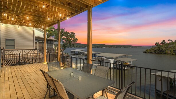 Une terrasse extérieure couverte, dotée d’une table et de chaises, qui offre une vue sur un plan d’eau et sur le coucher du soleil.