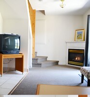Una sala de estar con televisión sobre un mueble de madera, una chimenea y una escalera que conduce a otra habitación.