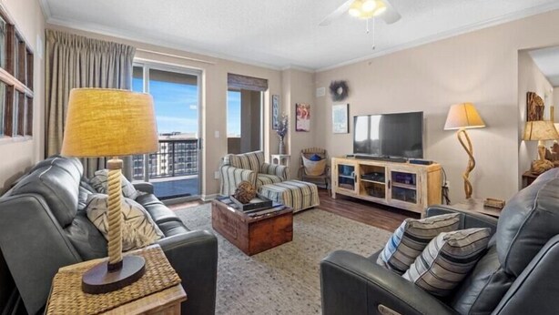 A living room with a sofa, coffee table, and TV. There is a balcony with curtains and a view of the sky.