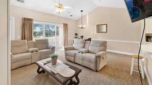 A living room with two beige sofas, a wooden coffee table, a ceiling fan, a large TV mounted on the wall, and a view of the outdoors through a sliding glass door. - Golf Villa Charm | Poolside Views | Relax in Style |  Magnolia North 4847-303 (Myrtle Beach)