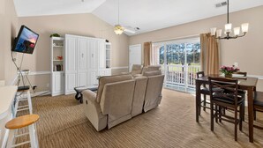 A living room with a beige sofa, a dining table with chairs, a TV mounted on the wall, and a ceiling fan. - Golf Villa Charm | Poolside Views | Relax in Style |  Magnolia North 4847-303 (Myrtle Beach)