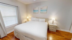 A neatly arranged bedroom with a large bed, two bedside tables with lamps, a window with blinds, and two wall-mounted paintings. - Cozy Golf Villa | Pool View Paradise | Relax | Magnolia North 4872-203 (Myrtle Beach)