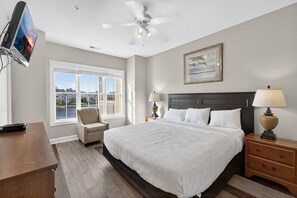 Quarto com cama grande, ventoinha no teto, TV na parede, uma cadeira, escrivaninha e janela com vista para a água.