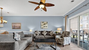 A modern living room with a grey sofa, a black coffee table, patterned armchairs, a ceiling fan, and artwork on the walls. - Pool View Bliss | Relax & Unwind | Myrtle Beach Awaits | Magnolia North 4827-202 (Myrtle Beach)