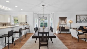 A spacious dining area with a long table and chairs, a kitchen with white cabinets, and a view of greenery outside. - Refreshed Magnolia Pointe Condo Near the Coast | Magnolia Pointe Luster Leaf 4855-101 (Myrtle Beach)