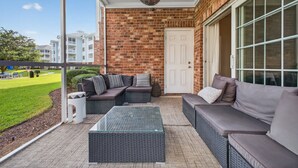 A covered patio with wicker furniture, a glass-top table, and a view of a brick building. - Refreshed Magnolia Pointe Condo Near the Coast | Magnolia Pointe Luster Leaf 4855-101 (Myrtle Beach)