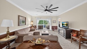 A living room with a sofa, coffee table, and TV. There is a ceiling fan and a window with curtains. - Spring Golf-View Rentals at Magnolia Pointe | Magnolia Pointe Luster Leaf 4878-201 (Myrtle Beach)
