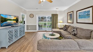 A living room with a television, a ceiling fan, a sofa, and a dining area with a view of the outdoors. - Tranquil Garden Oasis | Pool & Golf Views | Magnolia Pointe Luster Leaf 4882-104 (Myrtle Beach)