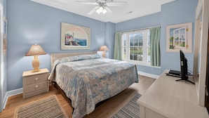 A bedroom with a bed, bedside lamps, a desk with a computer, a ceiling fan, and a window with curtains.