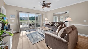 A spacious living room with a brown sofa, a coffee table, and a ceiling fan. There is a sliding glass door leading to a balcony with outdoor furniture.