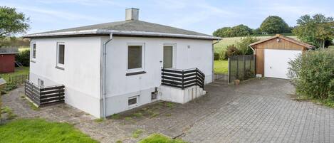En lille, etetages bygning med hvide vægge og et gråt tag. Den har en fordør, to vinduer og en lille veranda med gelænder i sort. Der er en belagt indkørsel, der fører til en garage med en trädør.