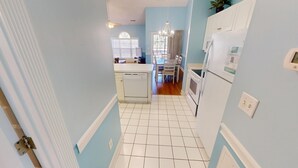 A narrow hallway with a kitchen area featuring white appliances, a dining table, and chairs.