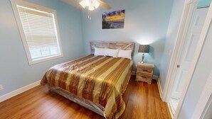 A bedroom with a wooden bed, a nightstand, a lamp, a painting on the wall, and a window with blinds.