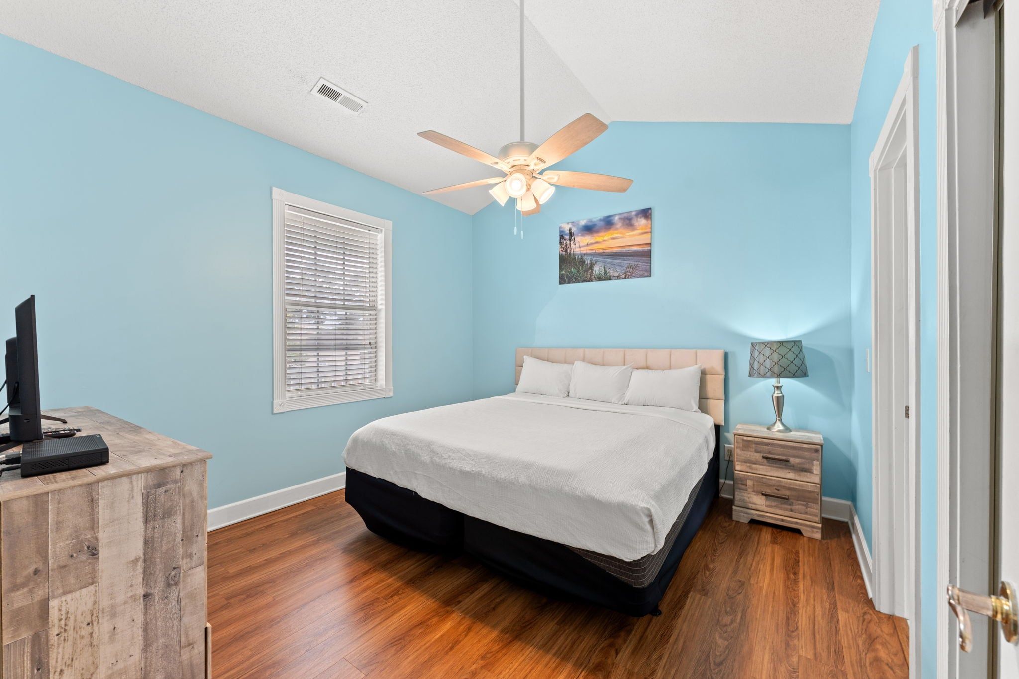 Quarto com cama, ventilador de teto, janela com persianas, mesa de cabeceira com abajur e escrivaninha com computador.