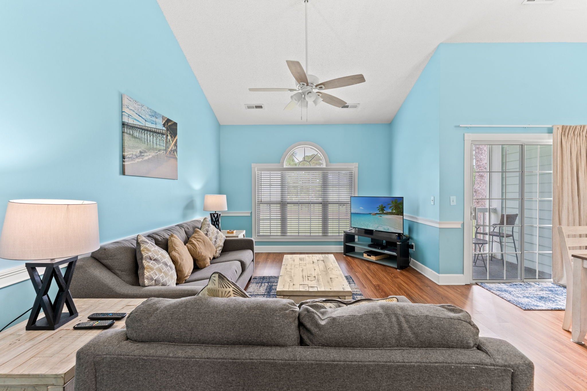 A living room with a sofa, a coffee table, a TV, a ceiling fan, and a window with blinds.