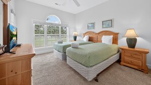 A bedroom with two beds, a ceiling fan, a desk, and a window with a view of greenery.